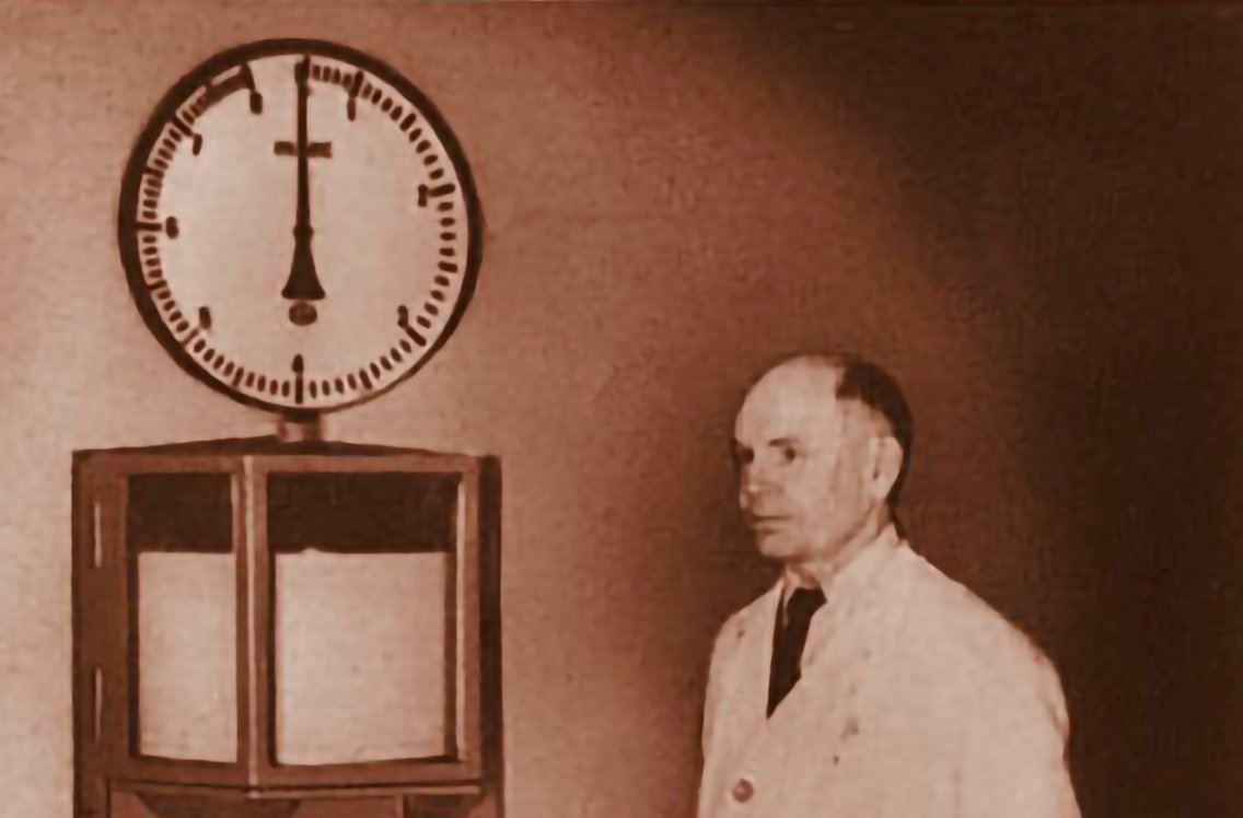 The image shows an old photo of an engineer in front of a mechanical measurement instrument
