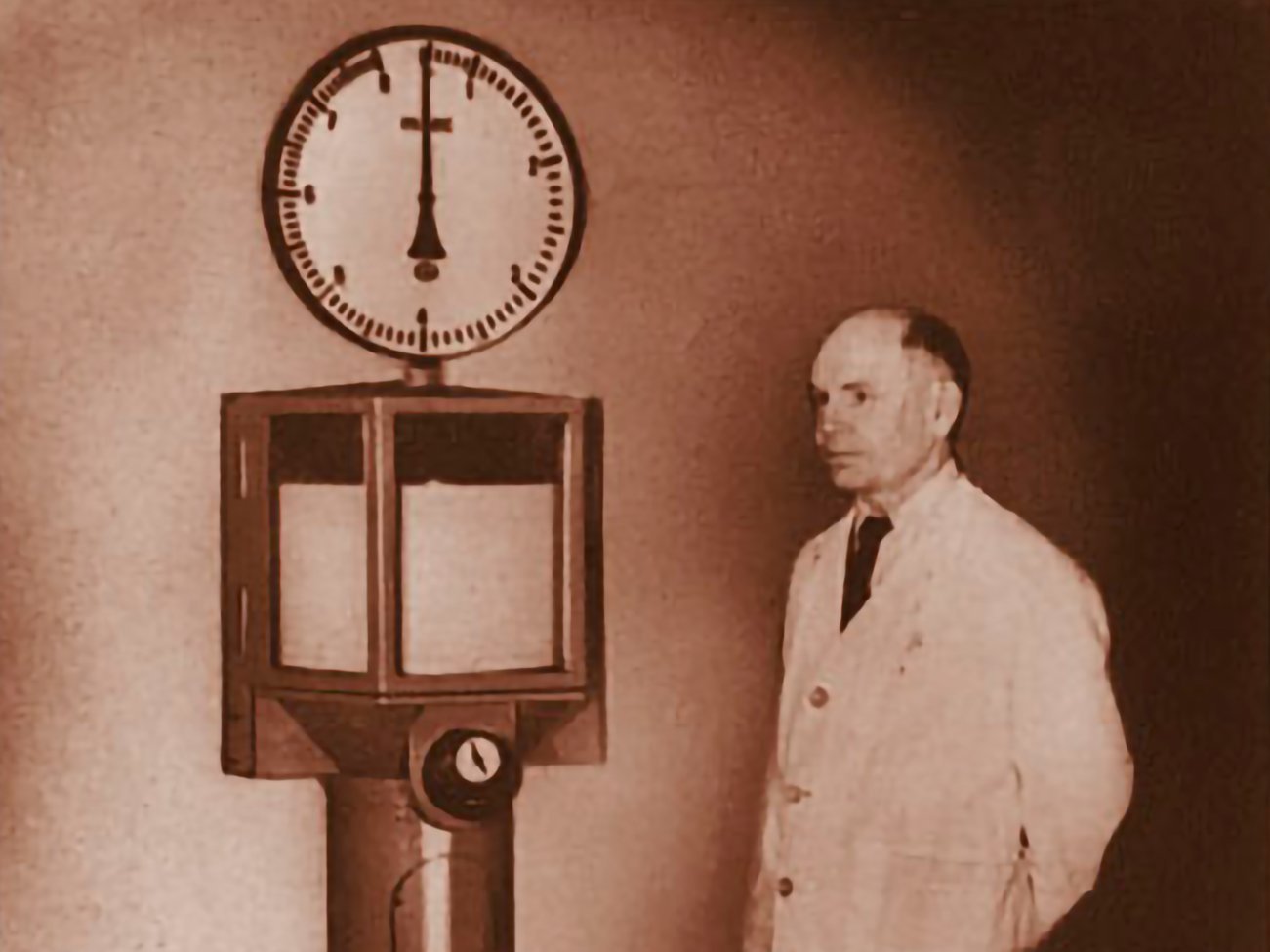 The image shows an old photo of an engineer in front of a mechanical measurement instrument