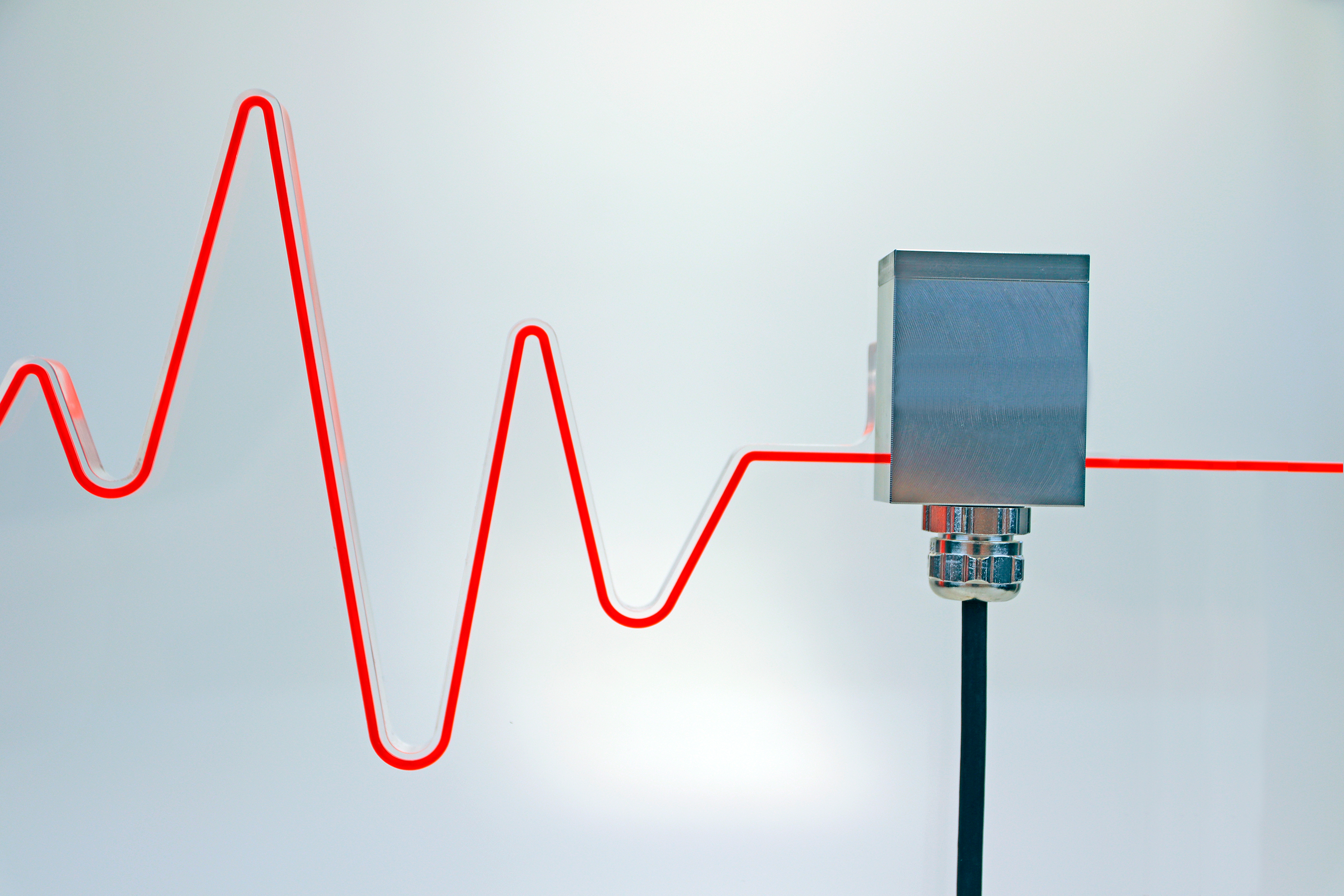 The picture shows an acceleration sensor in flange design - a red sinusoidal curve shape can be seen in the background, symbolising an acceleration force.