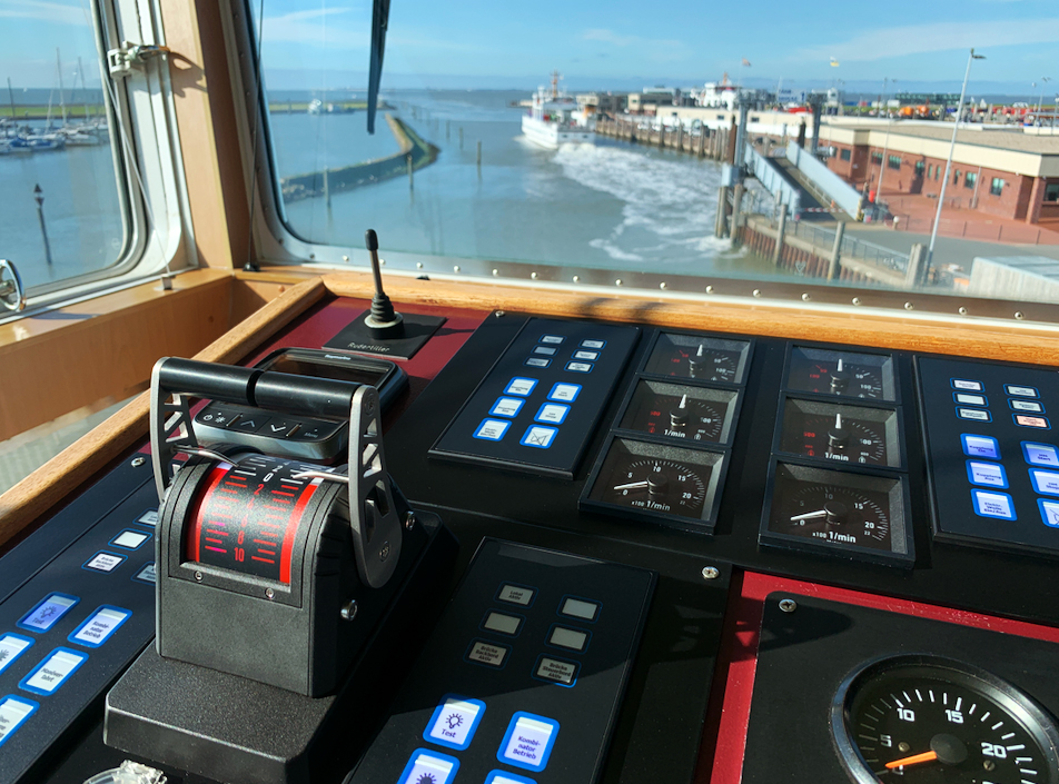 The image shows a bridge of a ship with a view over a bridge panel with control a lever and different control buttons and windwows in the background which show a habour scene