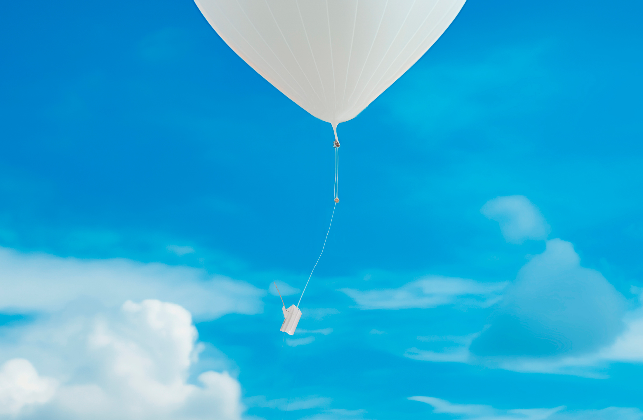 Das Bild zeigt einen weißen Ballon mit einer weißen Radiosonde. Im Hintergrund ist ein strahlend blauer Himmel mit einigen Wolken zu sehen.